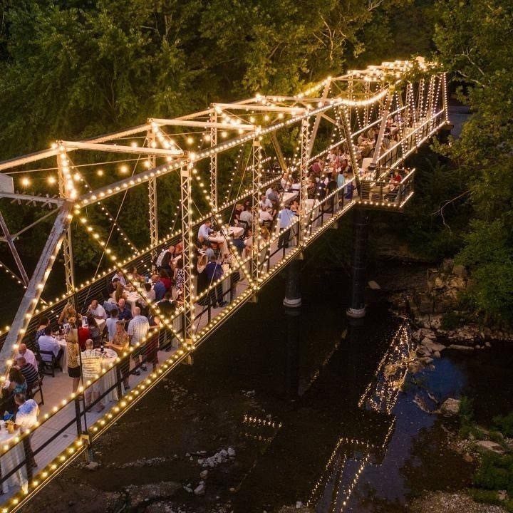 Illuminated metal bridge filled with wedding guests spanning a river in a forest.