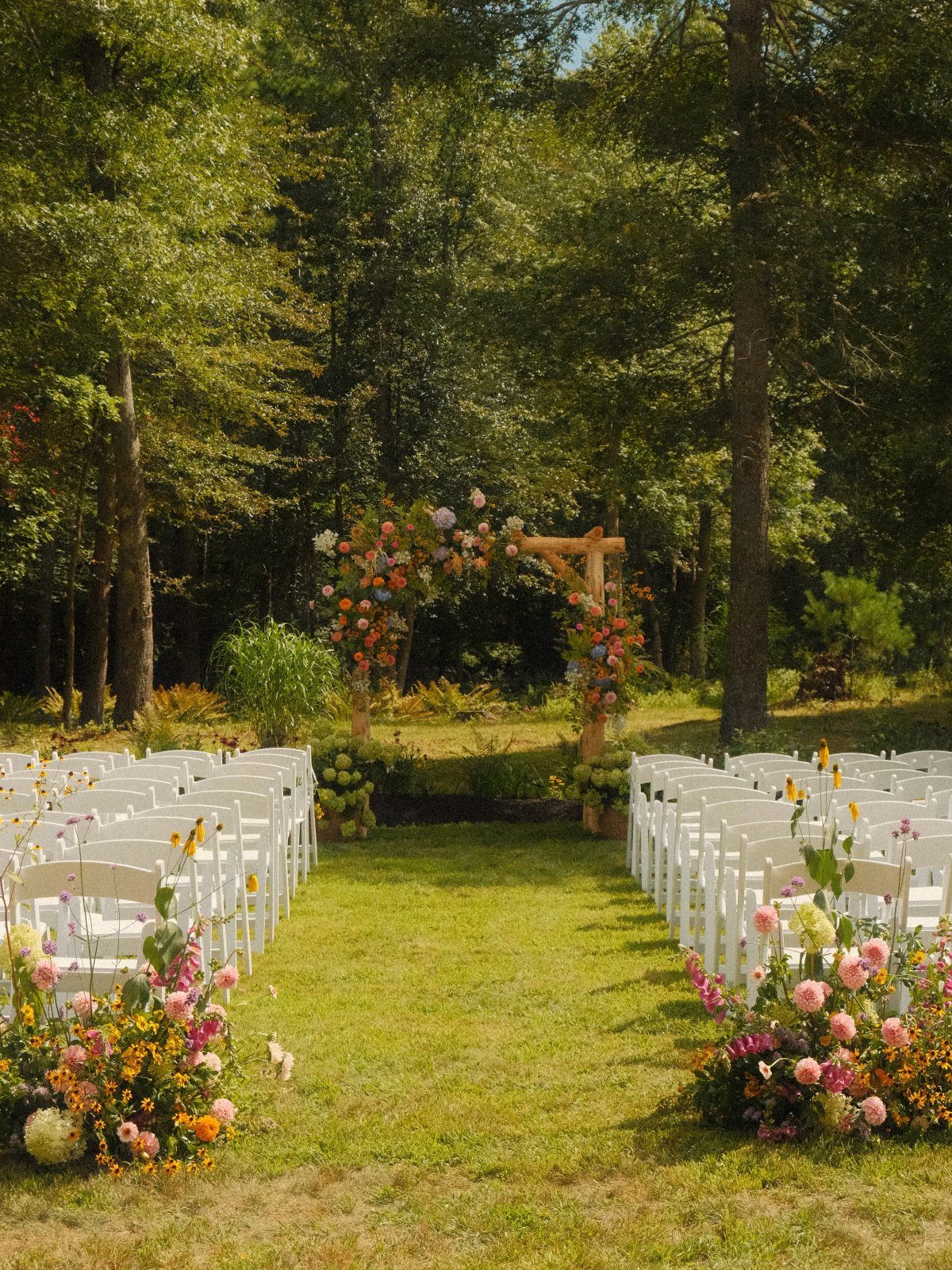 Outdoor wedding ceremony setup with white chairs and a floral wooden arbor in woods.