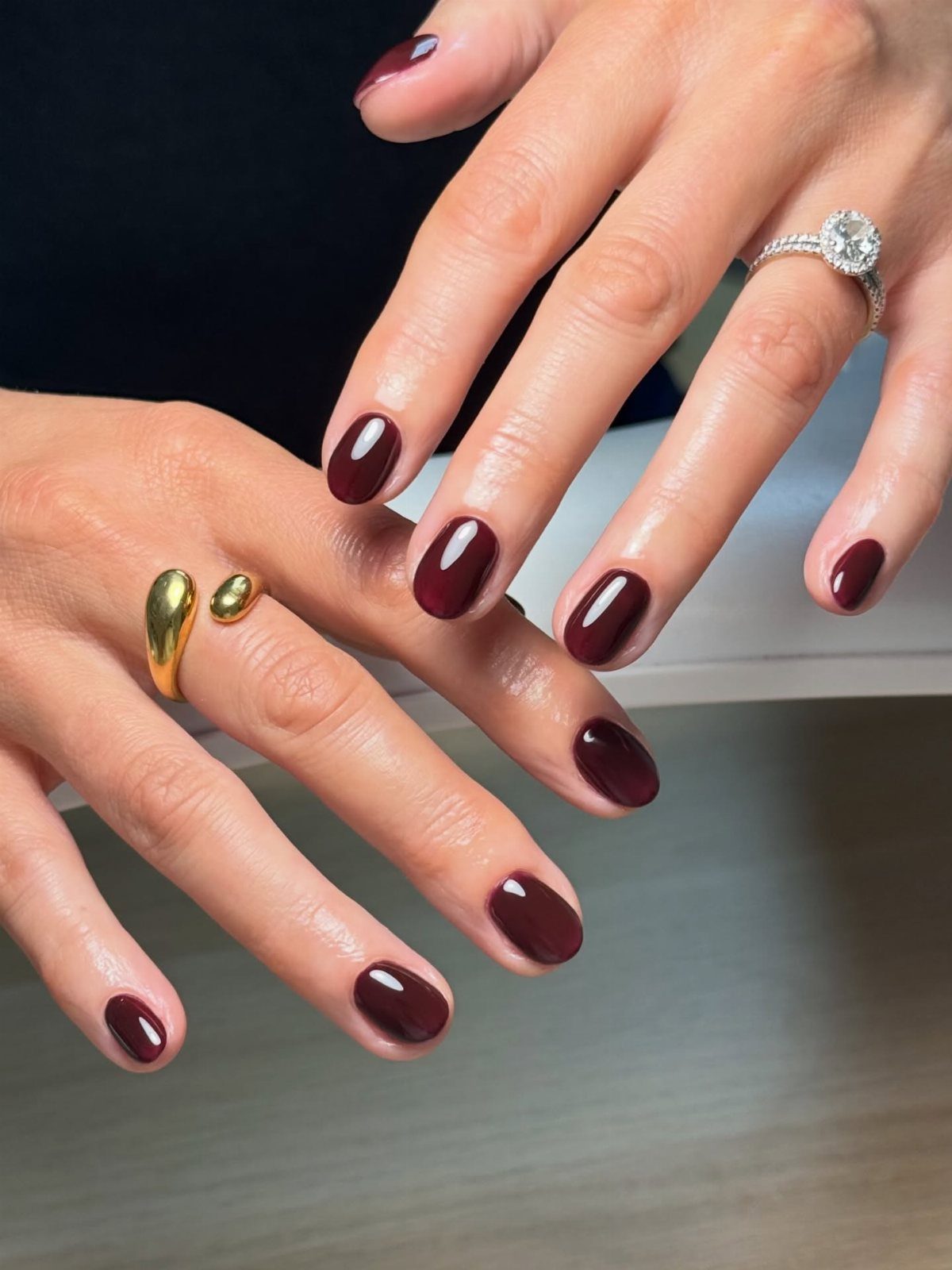 Hands with deep burgundy nail polish wearing a diamond engagement ring and gold rings.