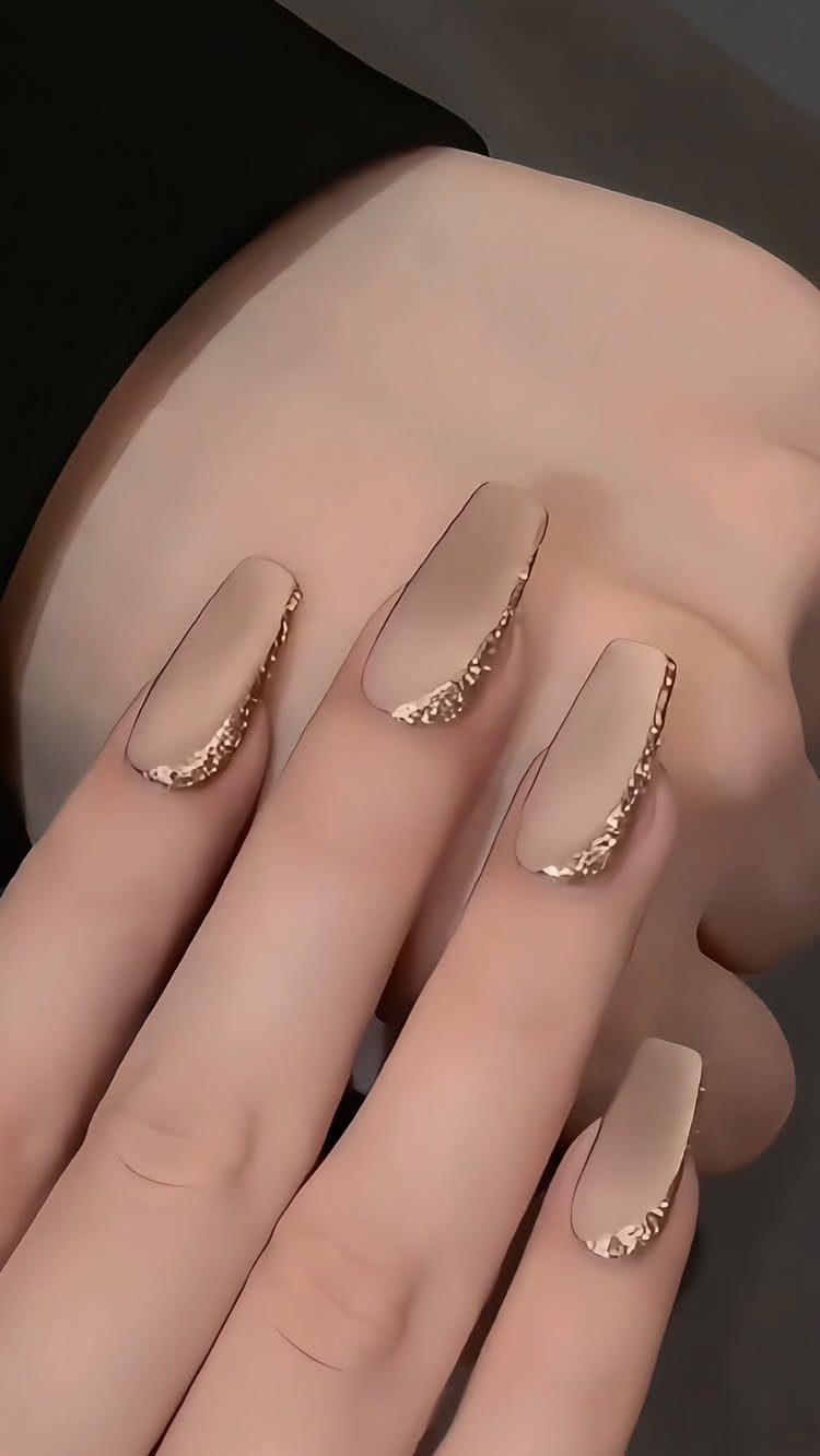 Nude coffin-shaped nails with gold glitter accents on a hand.