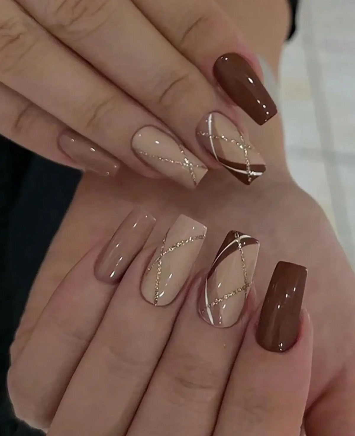 Long square nails with brown and nude polish and geometric glitter line art.