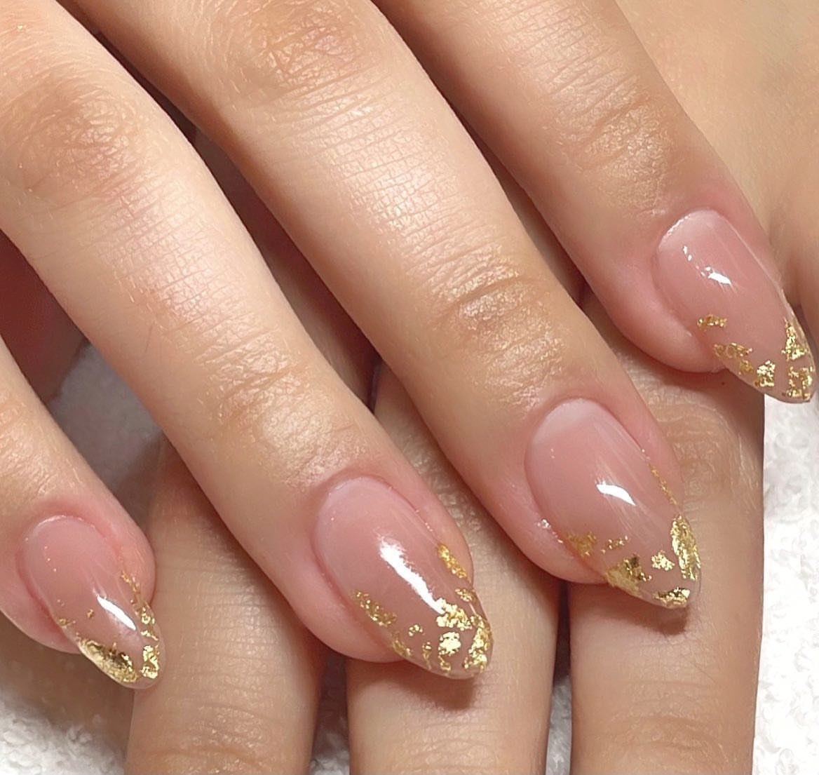 Almond-shaped nude pink nails with decorative gold foil flakes on the tips.