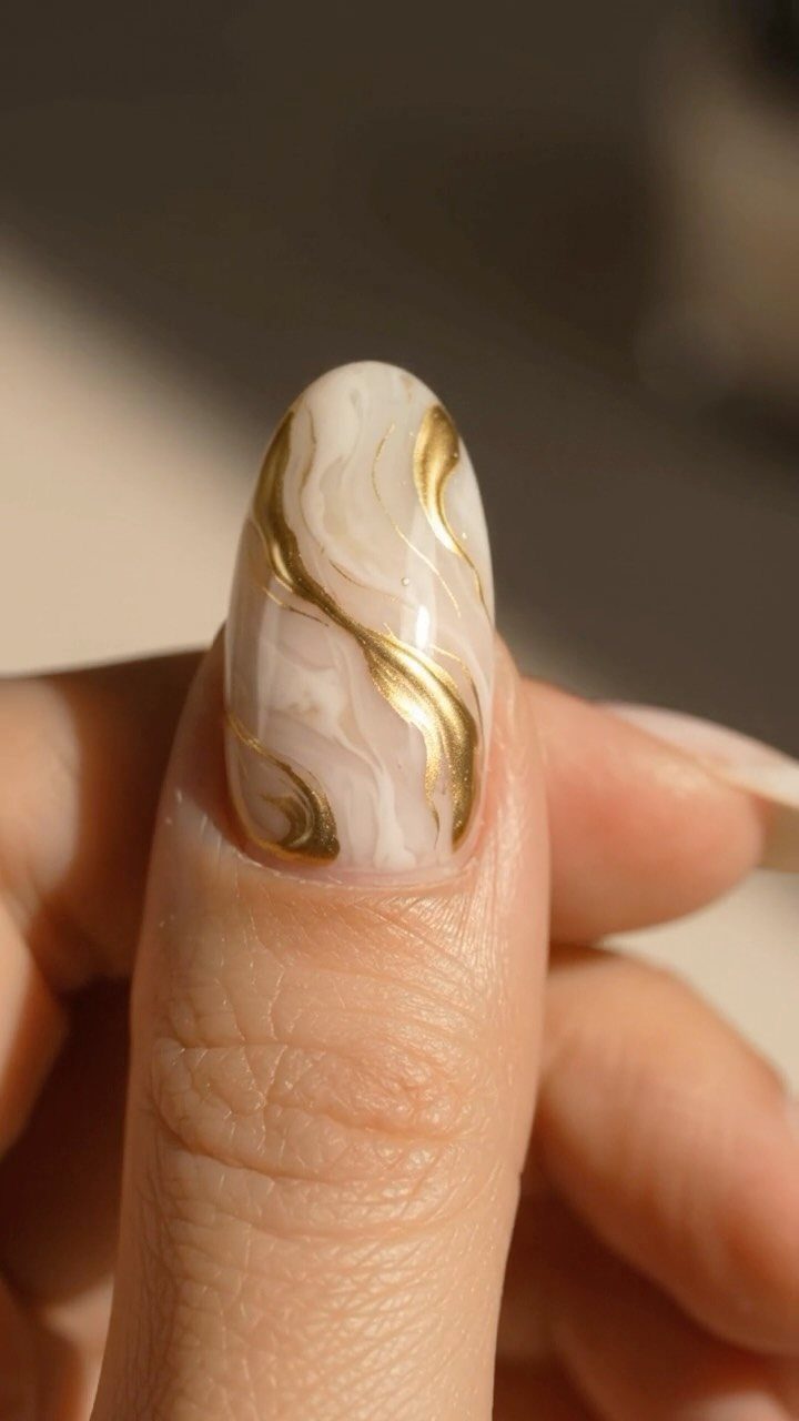 Oval fingernail with white and cream marble design and swirling gold metallic accents.