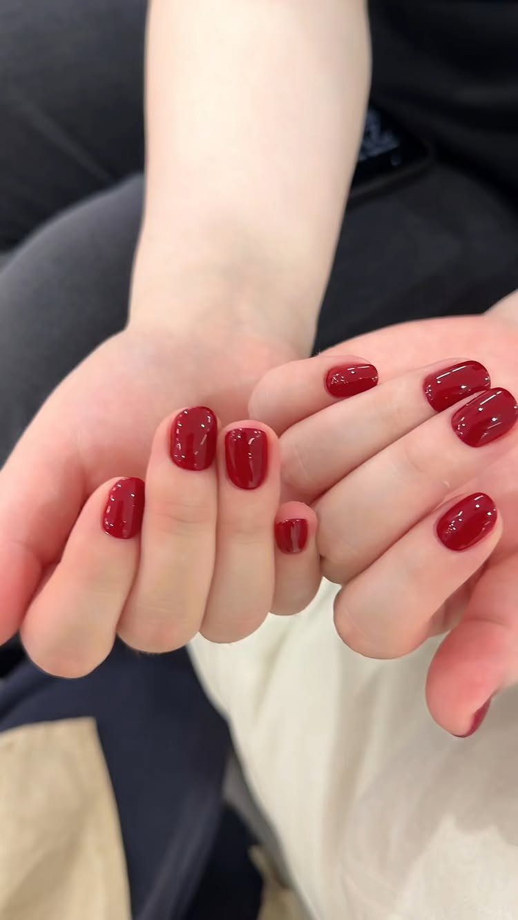 Short rounded nails painted in a glossy deep red polish on two hands.