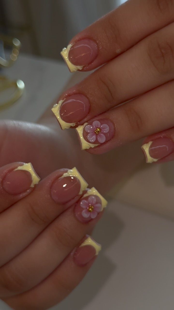 Almond nails with nude base, soft yellow French tips and small pink floral accents.