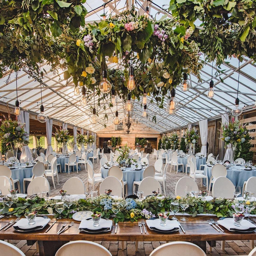 Glass greenhouse wedding venue with blue tablecloths, white chairs, and hanging greenery.