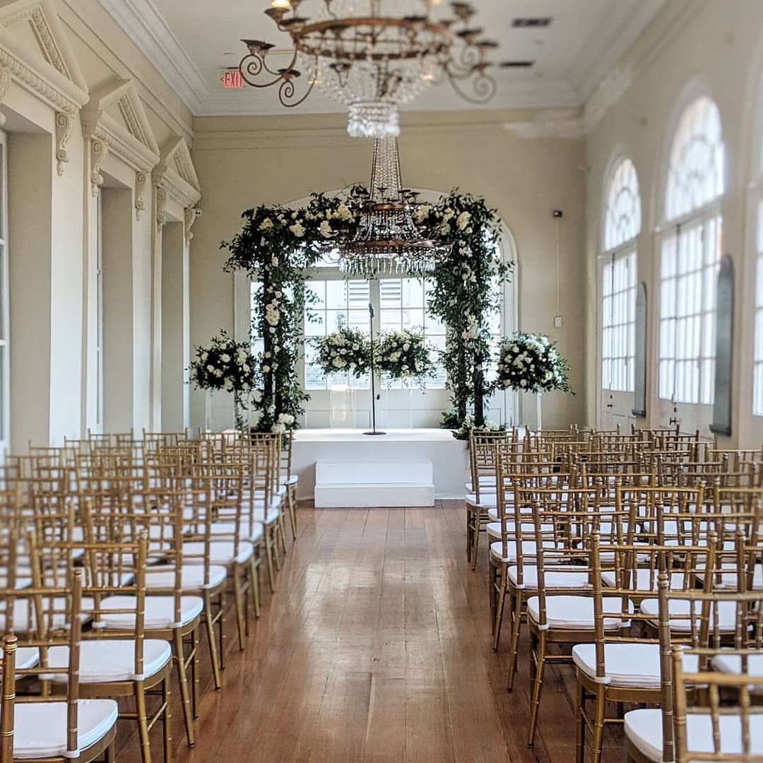 Gold wedding chairs facing a white altar with floral arches and crystal chandeliers.