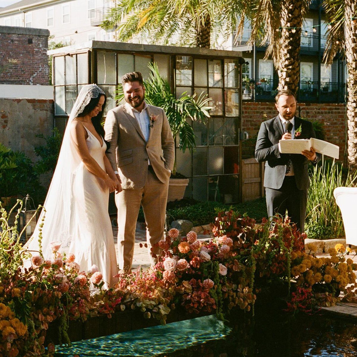 Bride and groom standing at an outdoor wedding ceremony with flowers and palm trees.