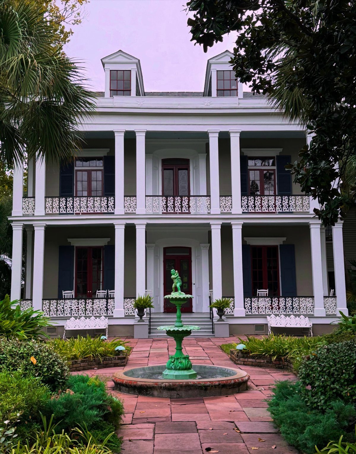 White historic two-story mansion with columns and a green tiered fountain in courtyard