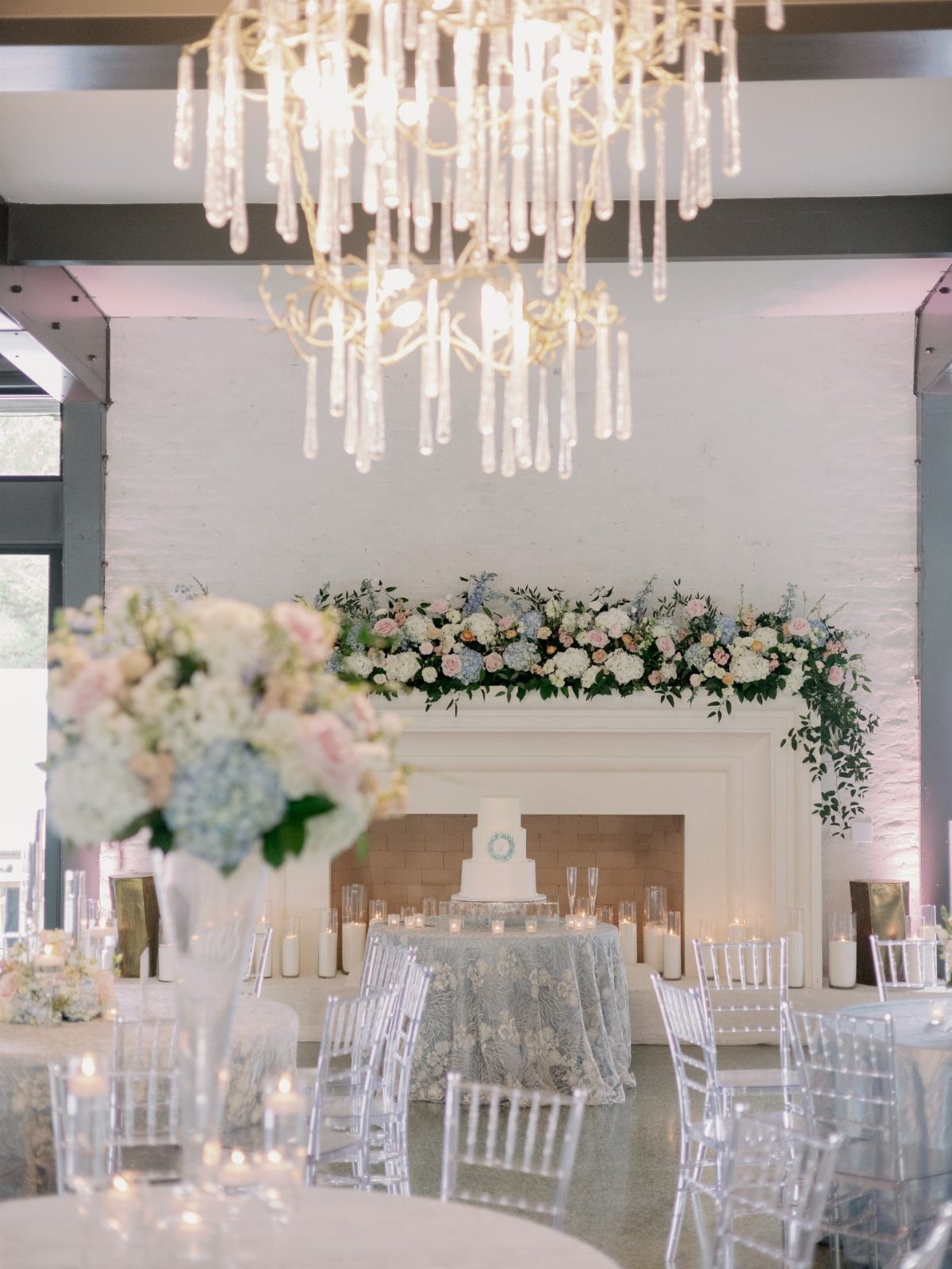 Wedding reception setup with a floral fireplace, white cake, and clear ghost chairs.