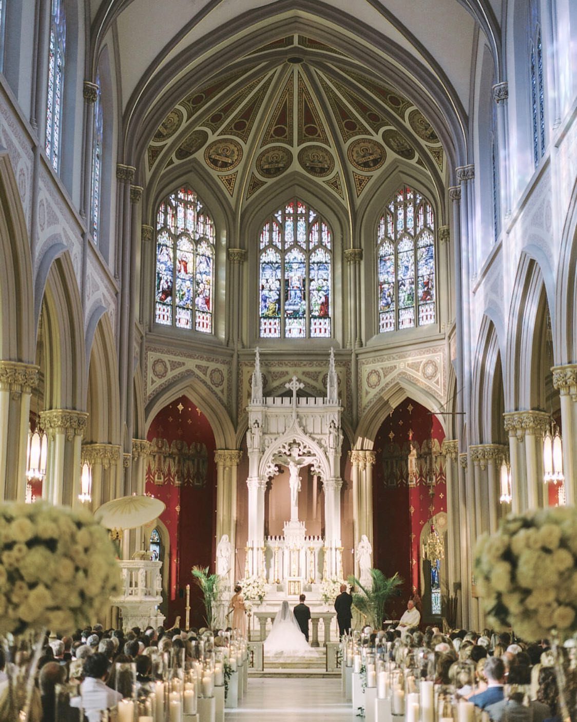 Wedding ceremony in a Gothic cathedral with stained glass and white floral arrangements.