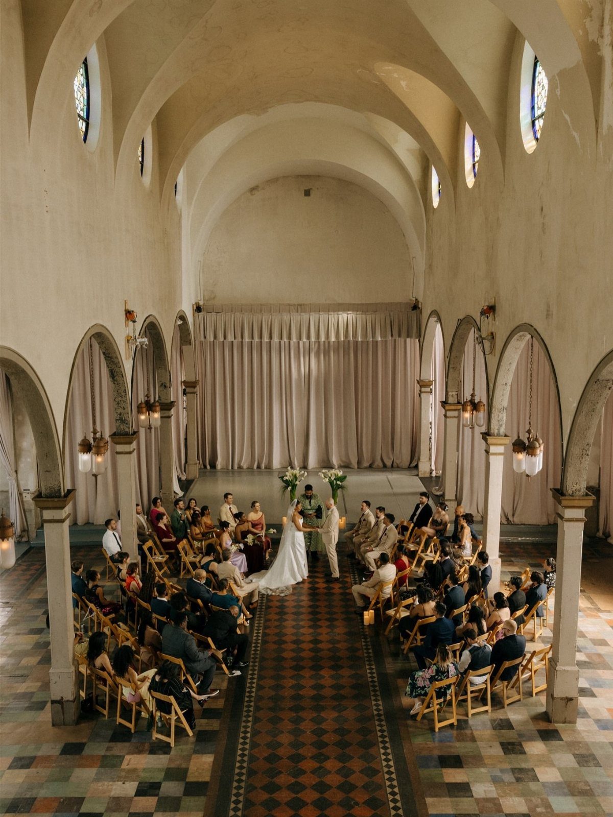 Wedding ceremony in a historic hall with vaulted ceilings and checkered tile floors.