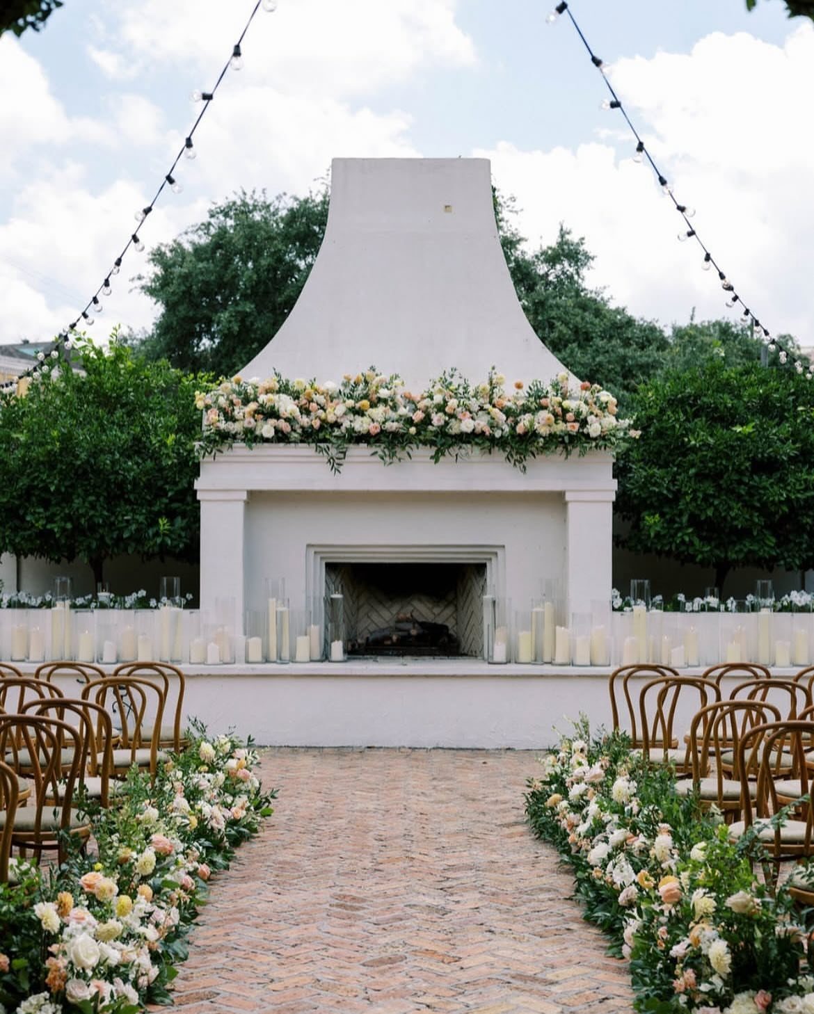 Outdoor wedding ceremony setup with a floral white fireplace and brick aisle.