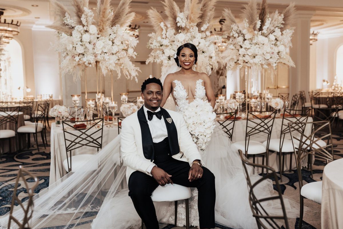 Bride and groom posing in a luxury ballroom with massive white floral centerpieces.