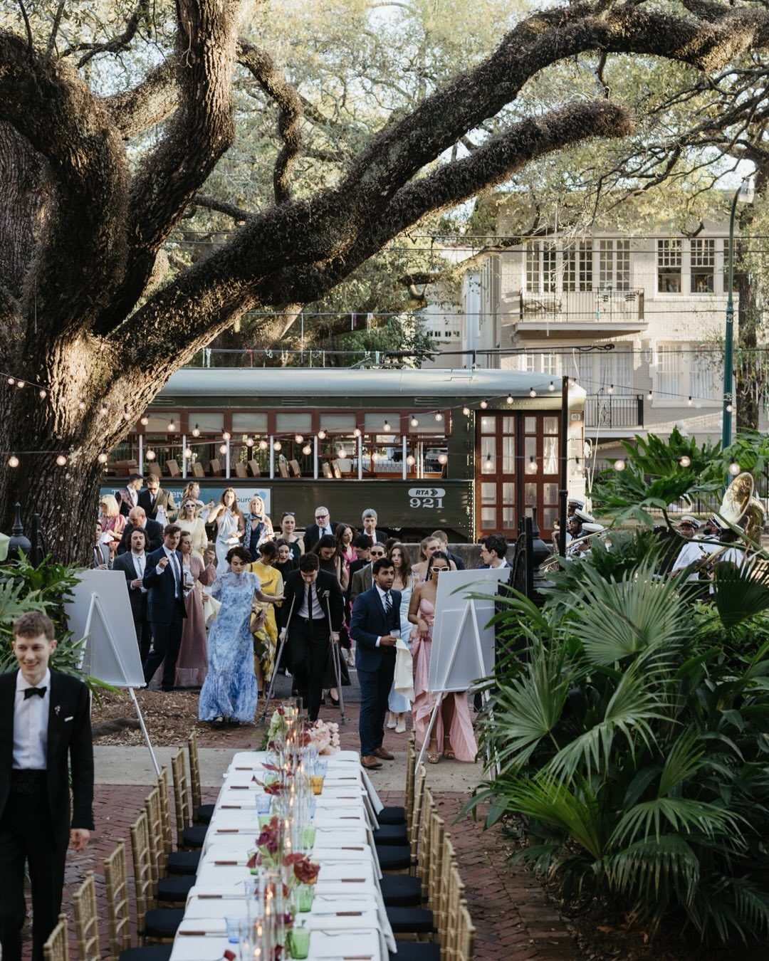 Outdoor wedding reception with a long white table and guests near a vintage streetcar.
