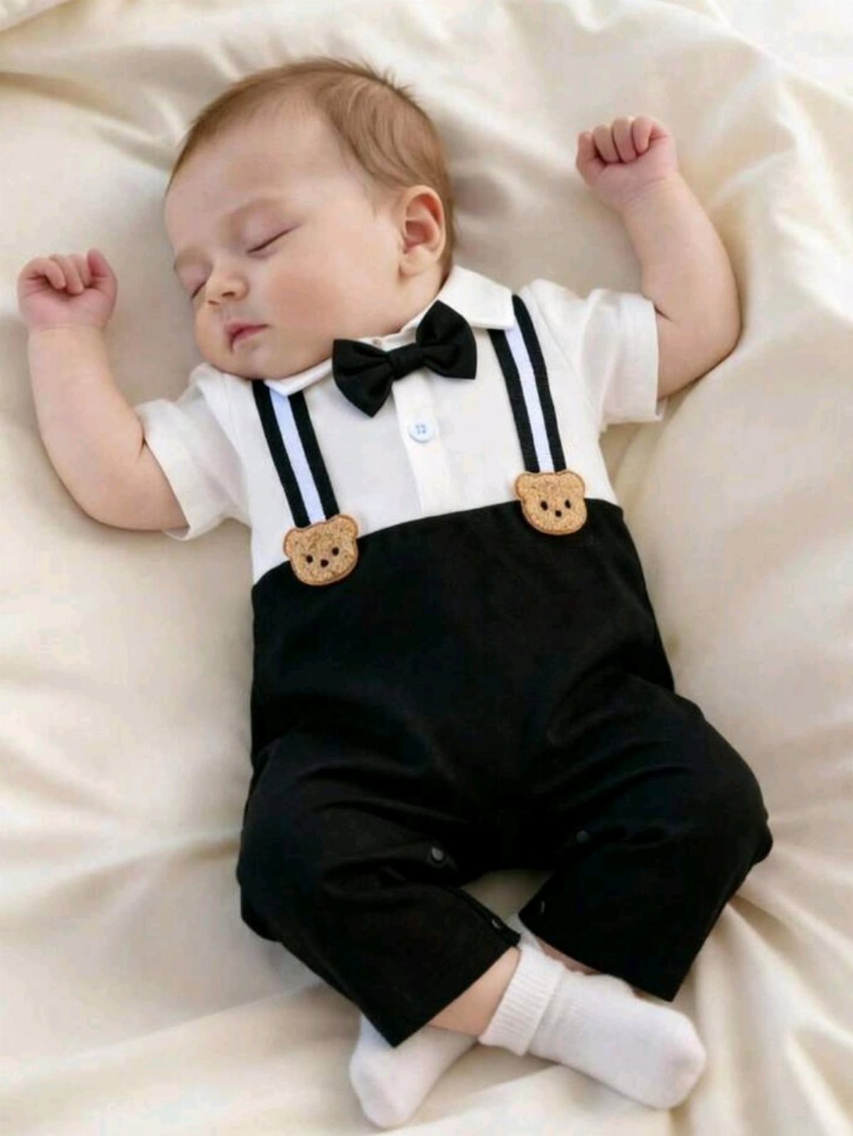 Sleeping newborn baby wearing a formal white shirt, black bow tie, and black overalls.