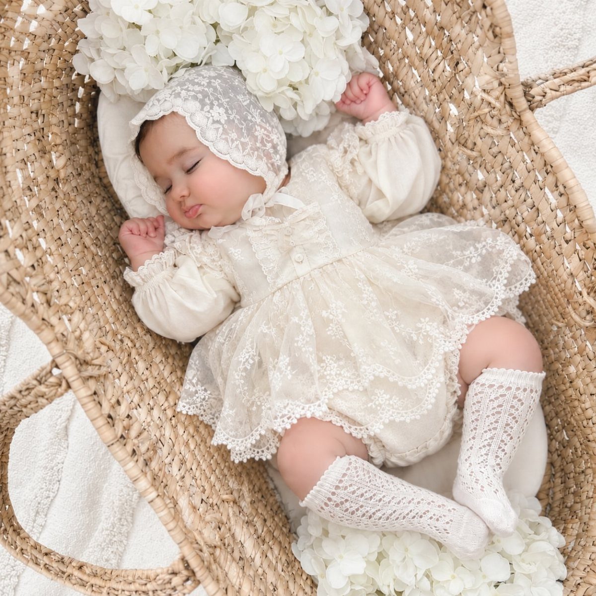 Sleeping newborn baby in a cream lace outfit and bonnet inside a wicker bassinet.