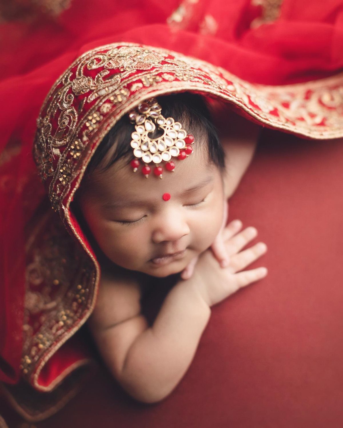 Sleeping newborn baby wearing traditional Indian gold jewelry and red embroidered fabric.