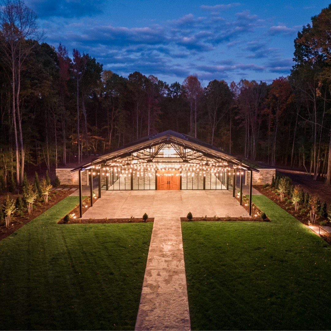 Modern rustic event pavilion with string lights and glass walls surrounded by forest trees.