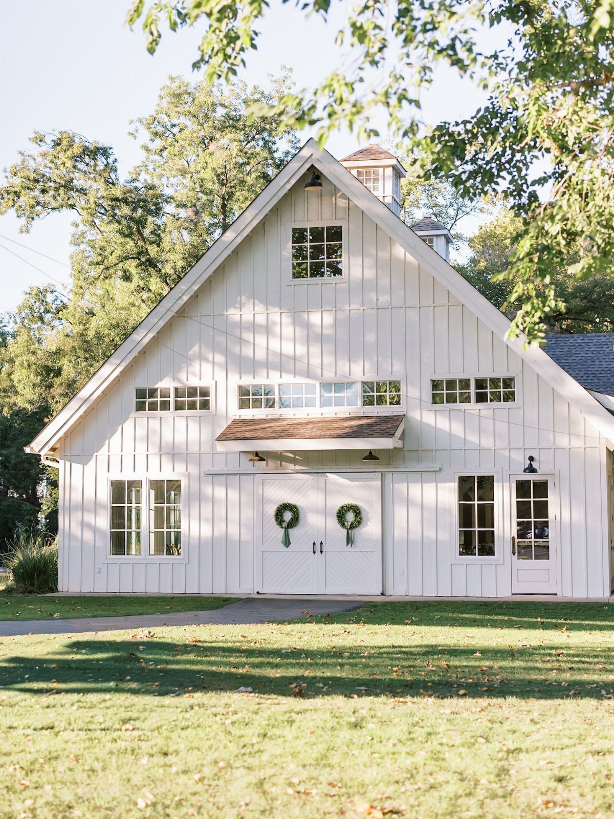 White wooden barn with green wreaths on doors set on a sunny lawn.