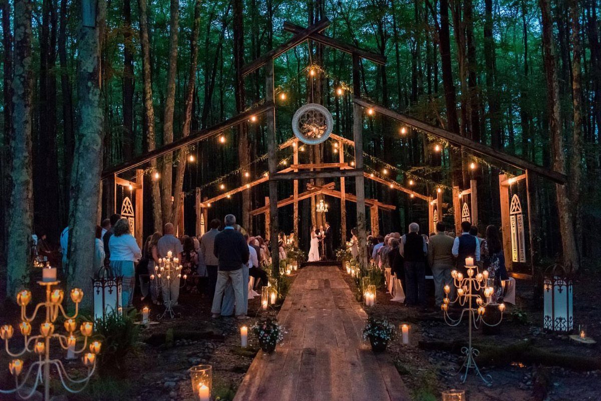 Rustic outdoor wedding ceremony with a wooden frame and string lights in a forest.
