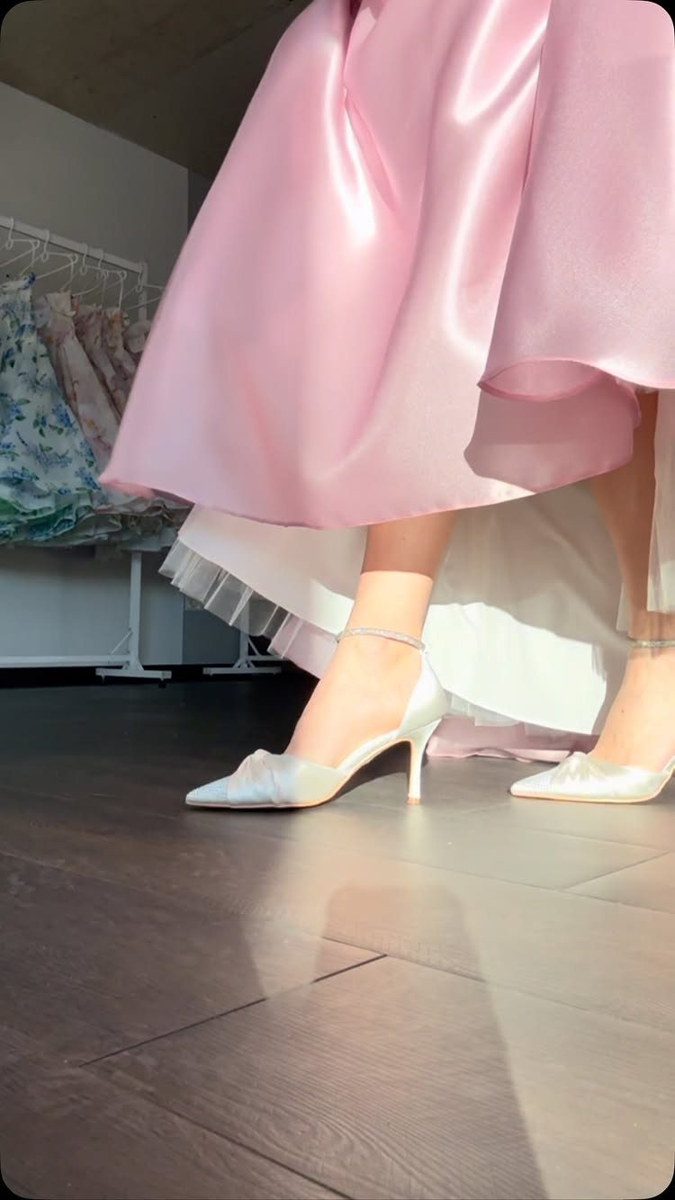 Pastel pink satin dress and white high heels on a dark wood floor.