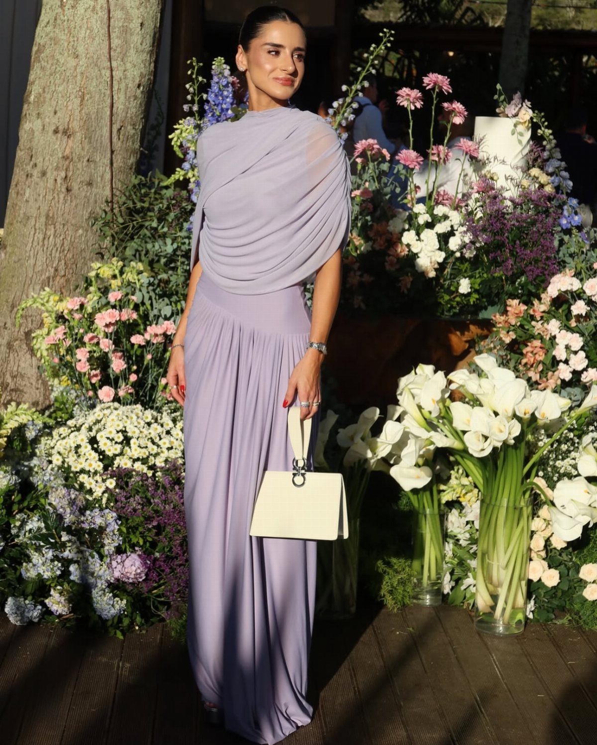 Woman in a draped lavender gown holding a white bag among floral arrangements.