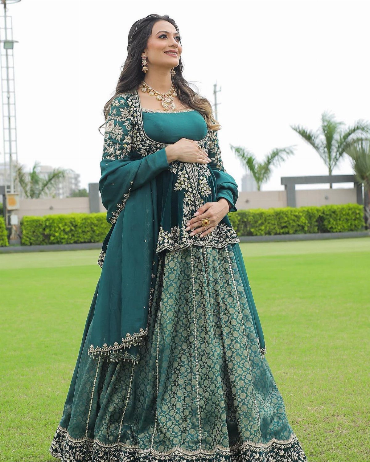 Pregnant woman in a teal embroidered lehenga and gold jewelry posing on a lawn.