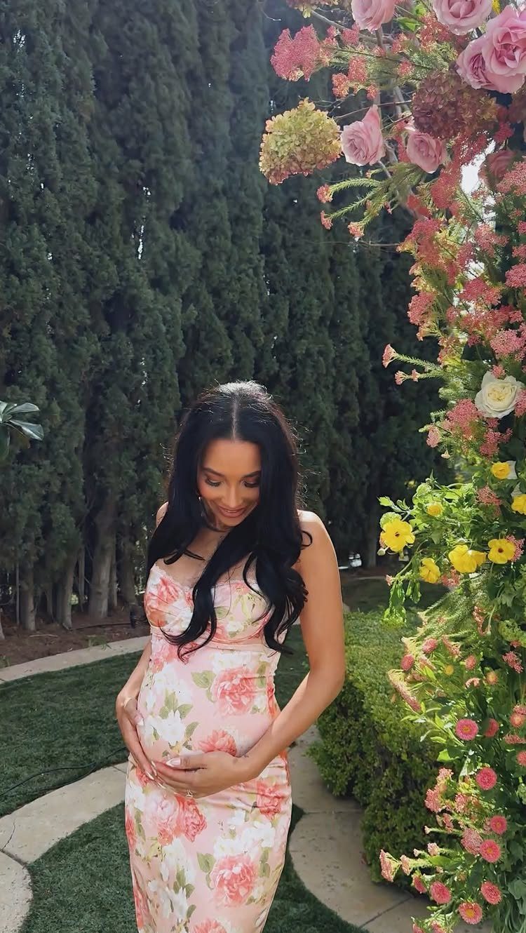 Pregnant woman in a pink floral dress standing by a colorful flower arch.