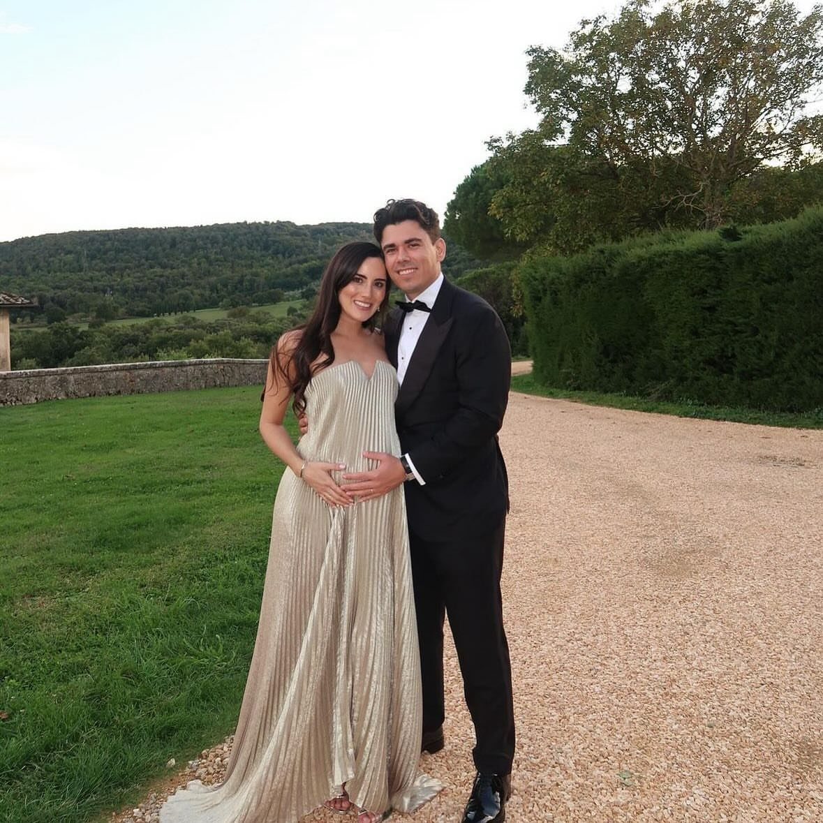 Pregnant woman in gold gown and man in tuxedo posing outdoors.