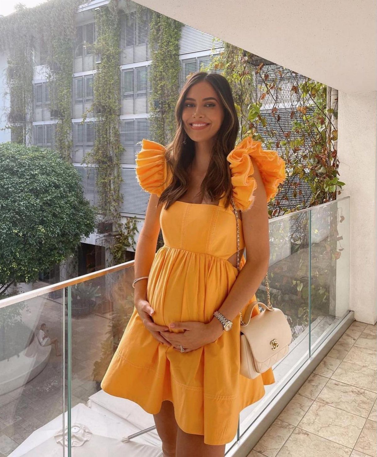 Pregnant woman in a short yellow ruffled dress posing on a sunny balcony