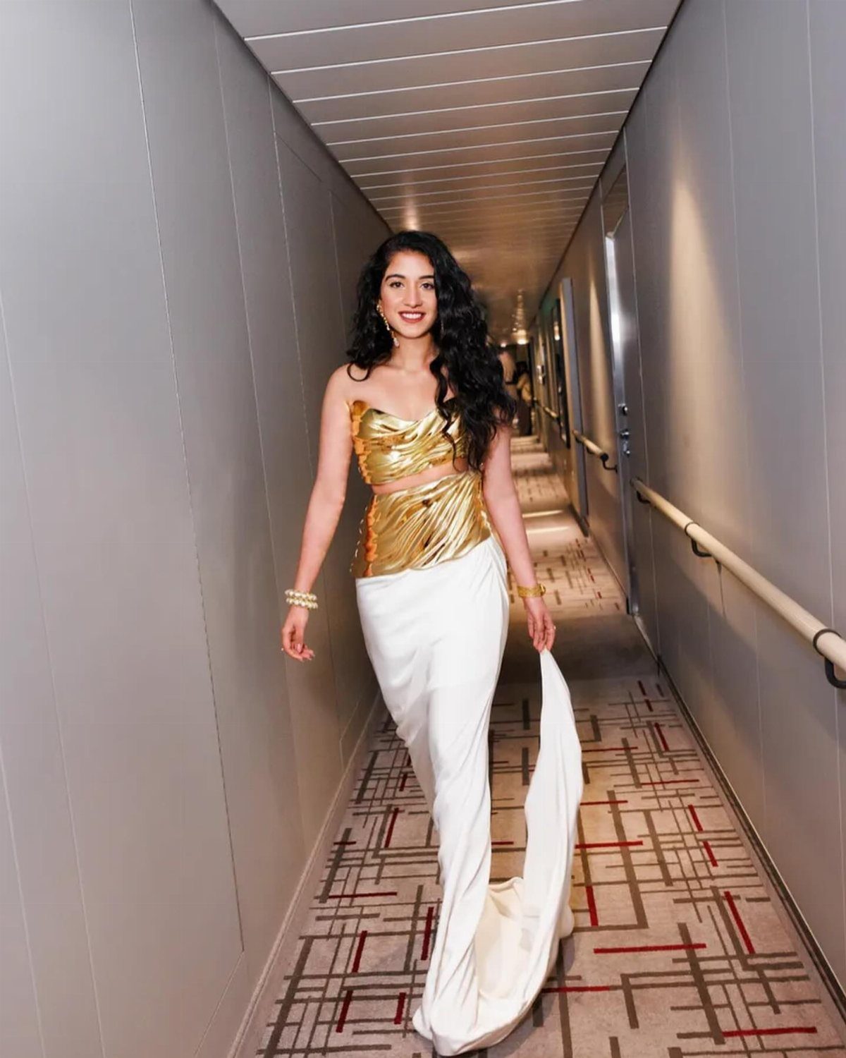 Woman in a gold metallic crop top and white skirt posing in a hallway