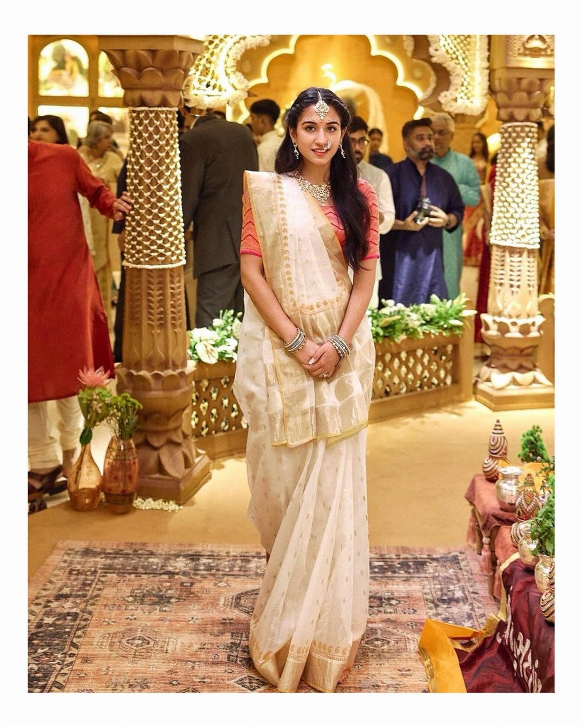 Woman in a white saree and red blouse posing in an ornate gold hall.