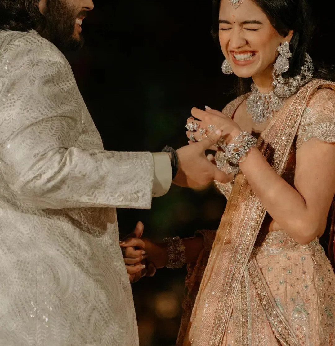 Woman in peach embellished lehenga and diamond jewelry laughing with a man.