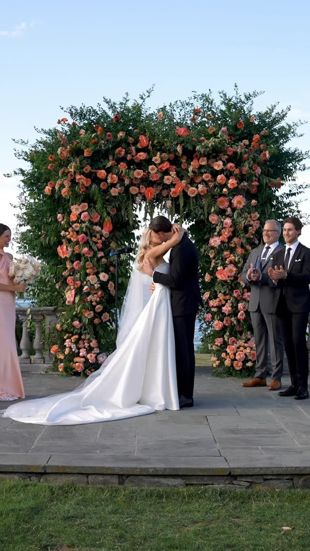 Bride and groom kissing under a lush floral arch on a stone patio.
