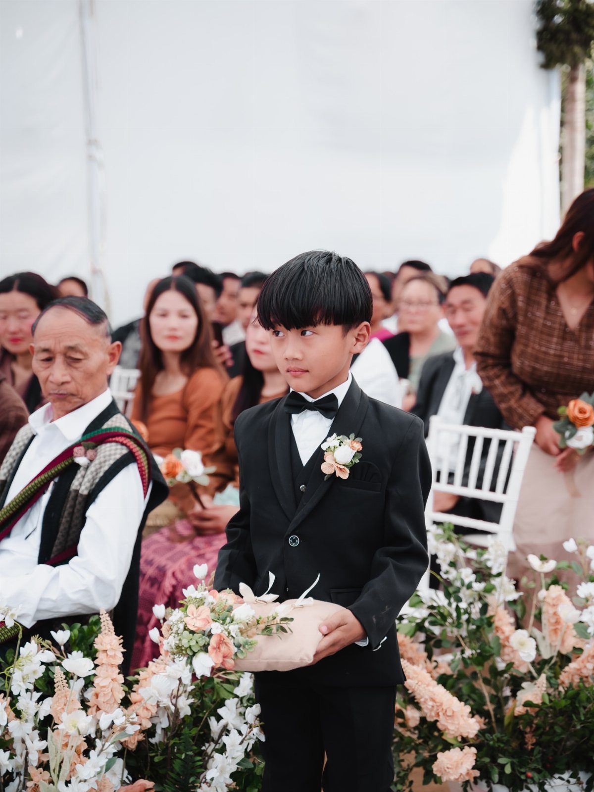 Young boy in a black tuxedo holding a flower pillow at a wedding