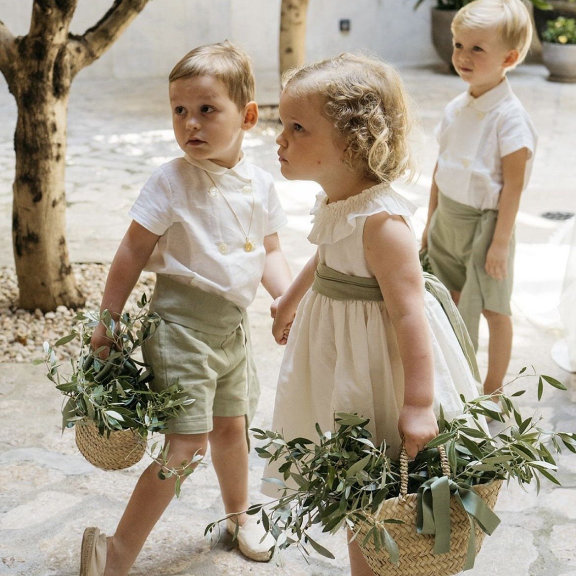 Children in white and sage green wedding outfits carrying baskets of olive branches.