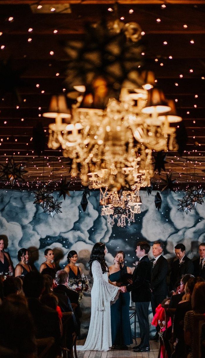 Wedding couple standing before a starry night backdrop under a large golden chandelier.