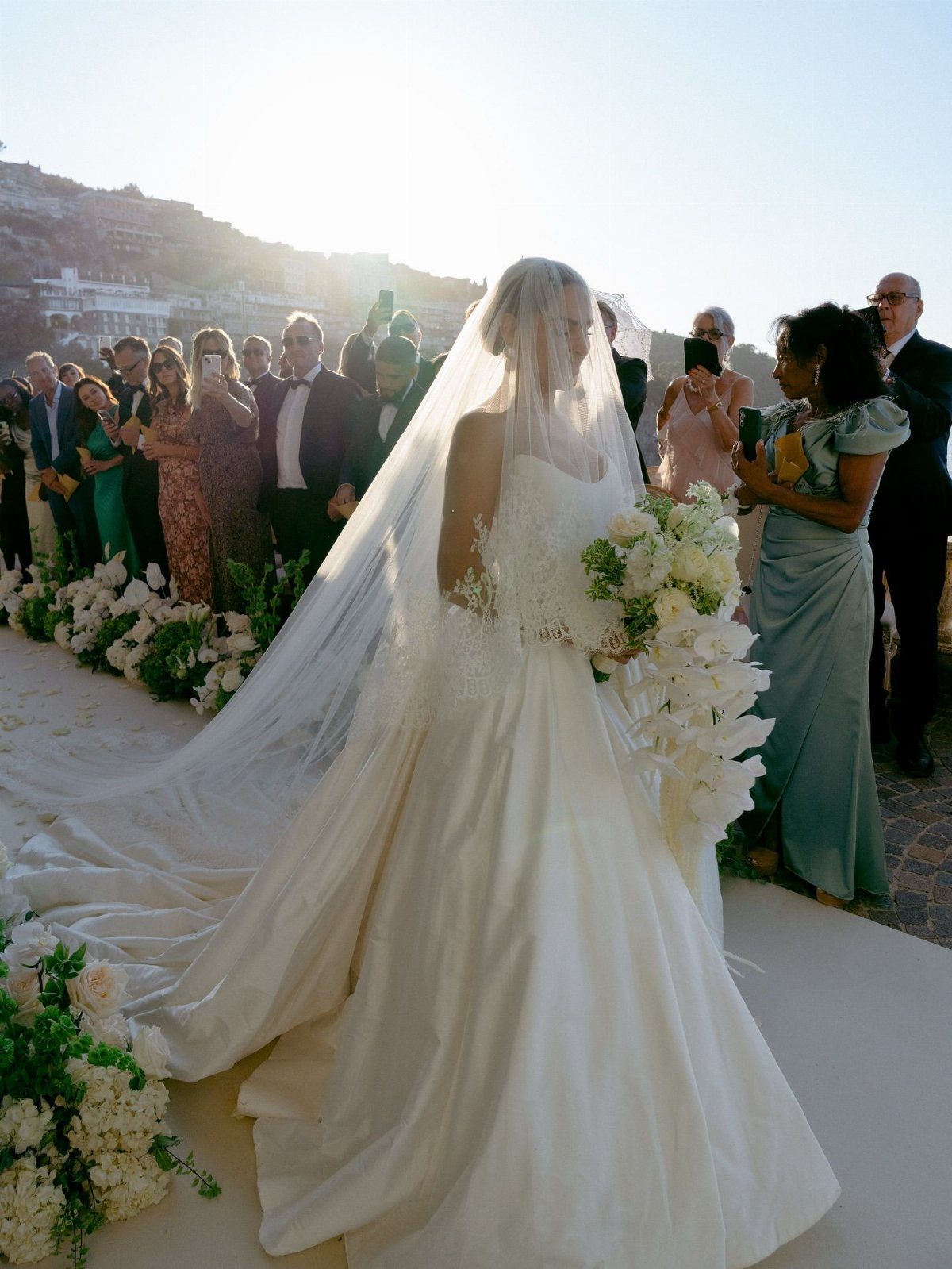 Bride in a white satin gown and veil walking down a flower-lined aisle.