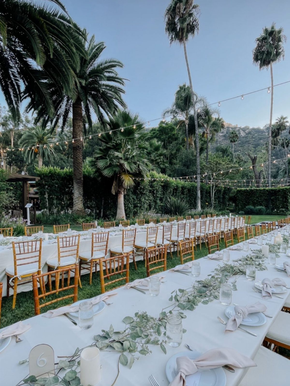Long white wedding tables with gold chairs in a lush tropical garden setting.