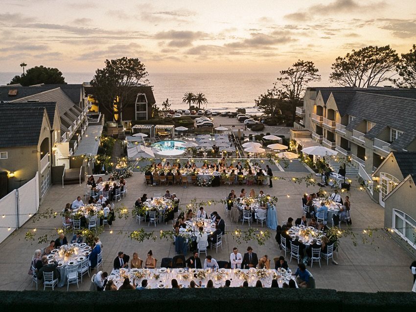Outdoor wedding reception with guests at white tables in a coastal resort courtyard at sunset.