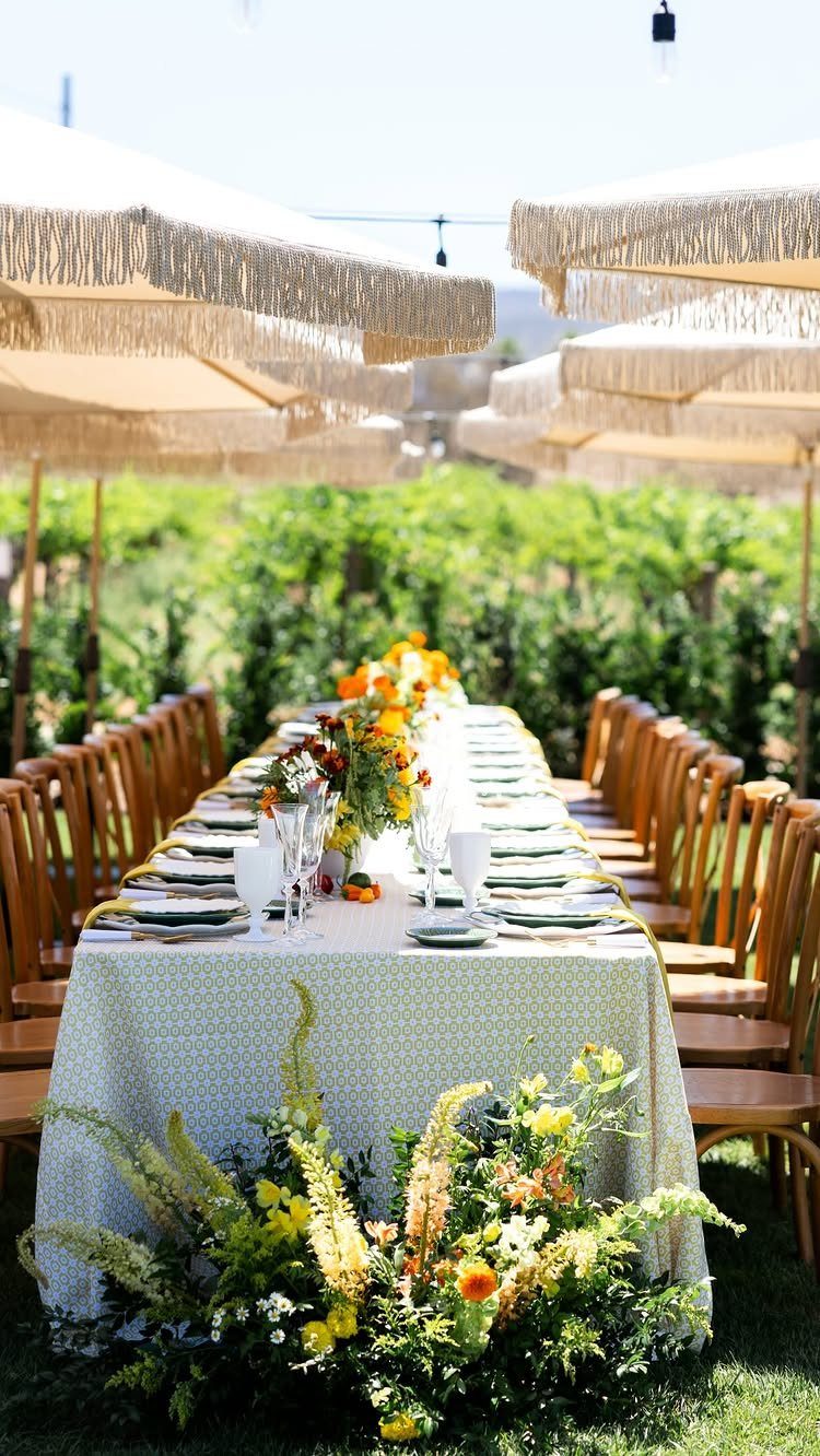 Long wedding table with yellow flowers and fringed umbrellas in a vineyard setting.
