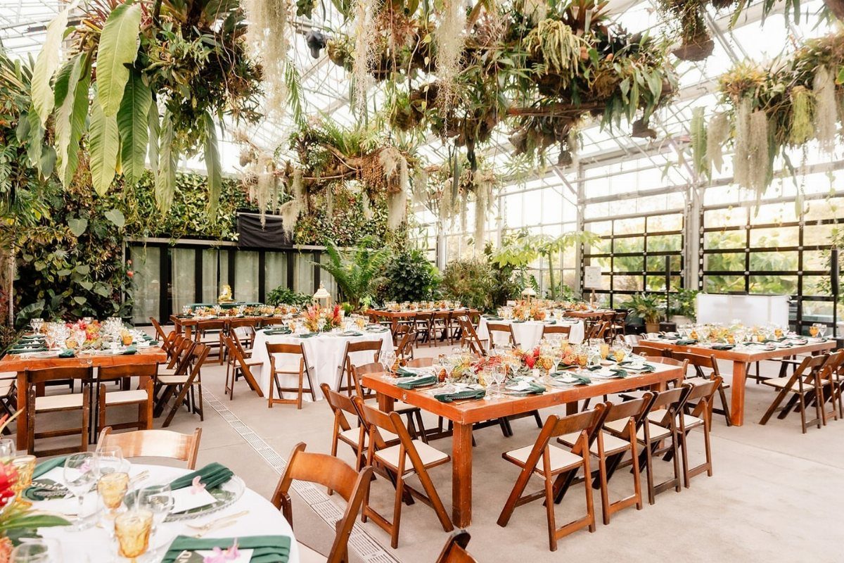 Wedding reception tables set inside a sunlit glass greenhouse with hanging tropical plants.