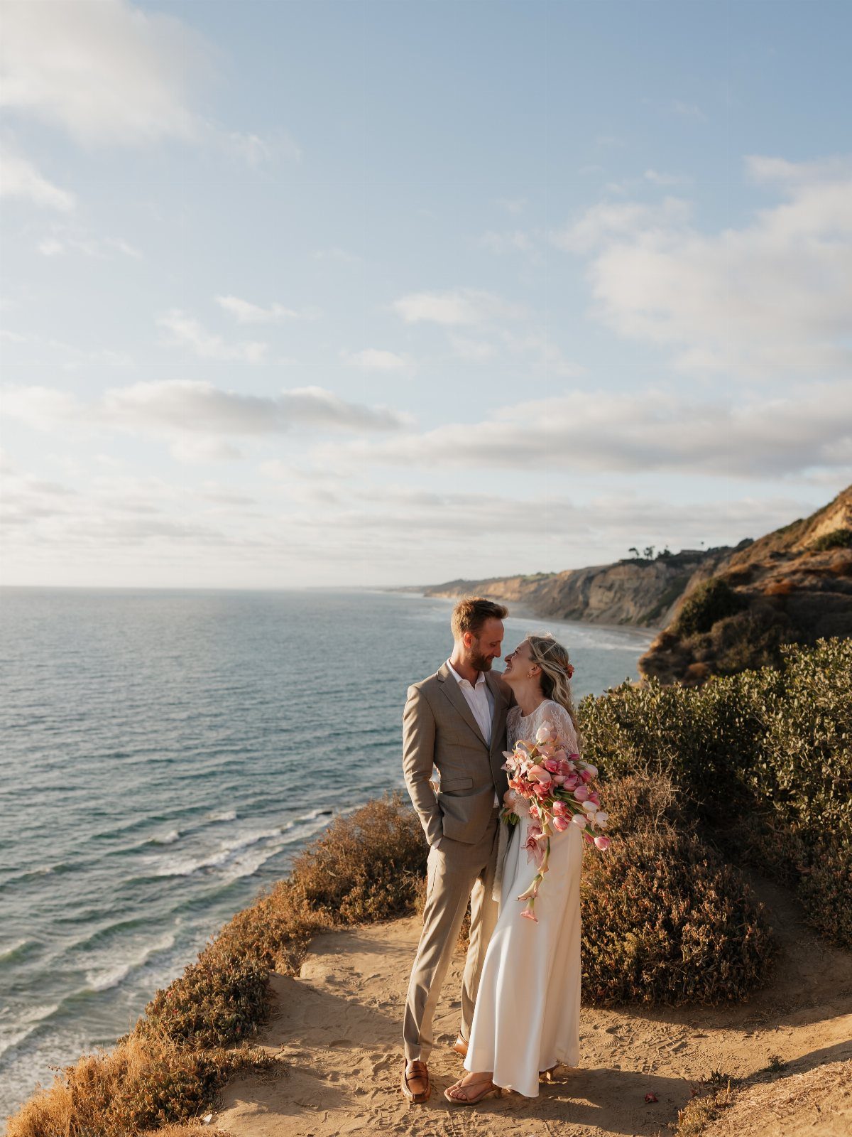 Bride and groom posing on a sandy cliff overlooking the blue ocean coastline.