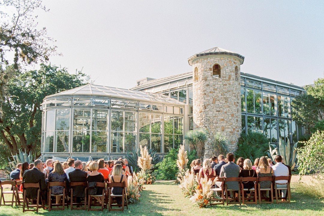 Wedding guests seated on a lawn before a large glass greenhouse with stone tower.