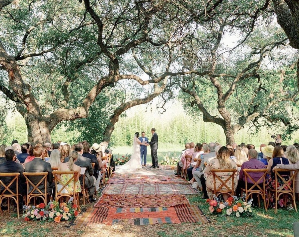 Wedding ceremony under large oak trees with guests seated along a rug aisle.