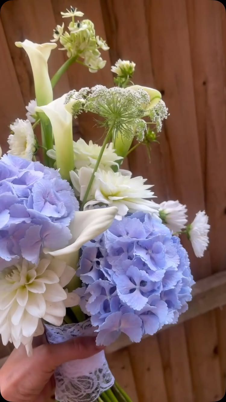 Bridal bouquet with blue hydrangeas, white calla lilies, and white dahlias wrapped in lace.