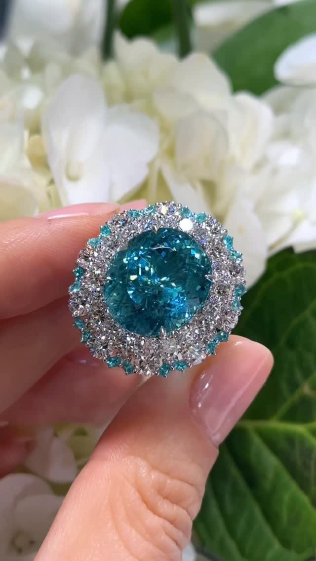Blue gemstone ring with diamond halo held against white flowers and green leaves.