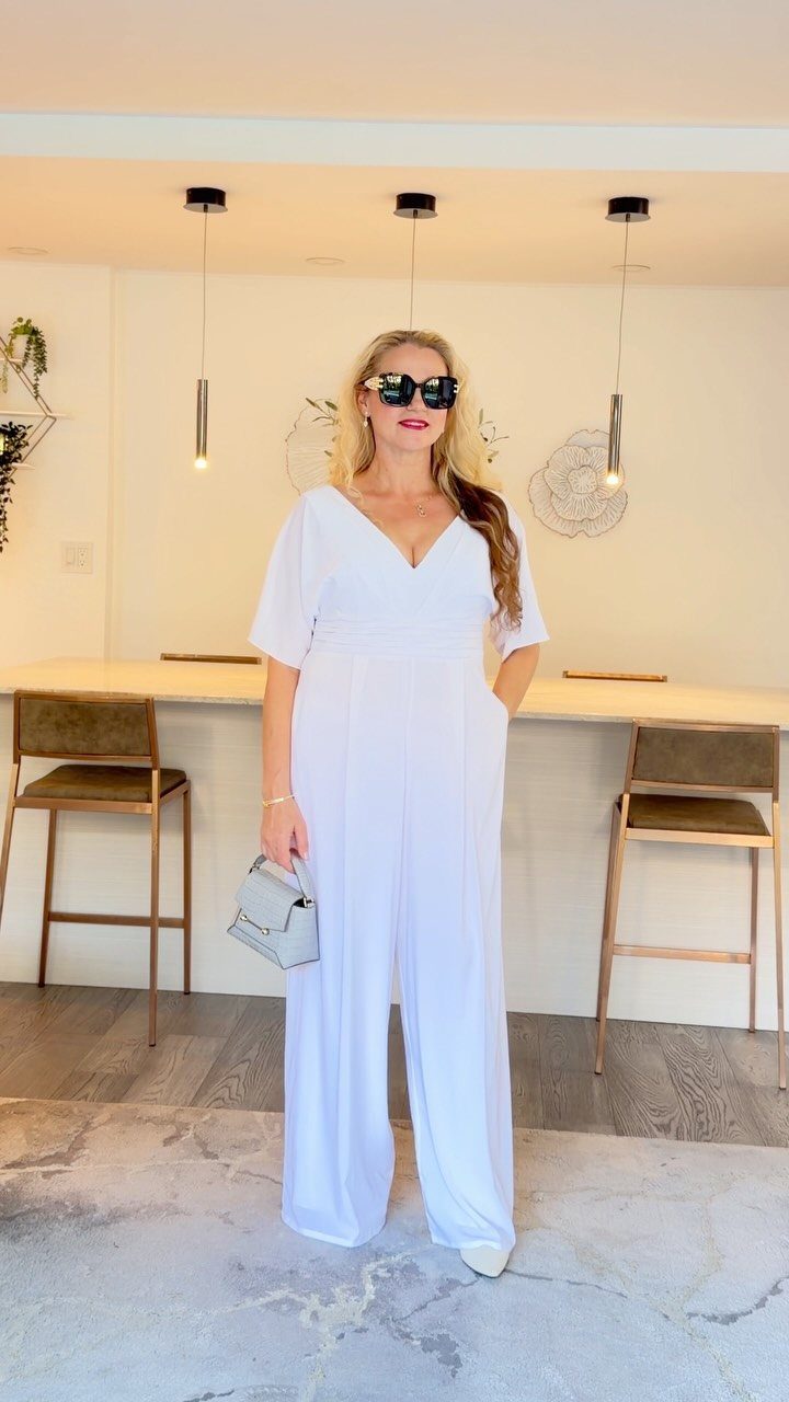 Woman wearing a white wide-leg jumpsuit and sunglasses in a modern kitchen setting.