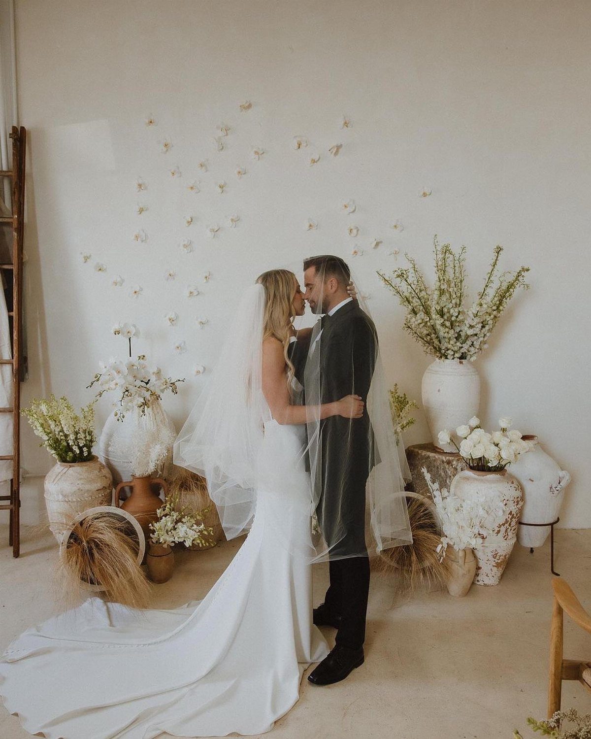 Bride in white wedding gown and groom in dark suit embracing in minimalist room.